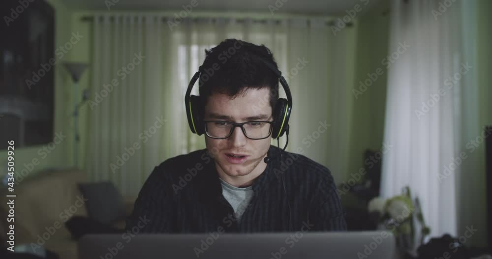 Portrait of male freelancer speaking to group chat via headset during remote work at home. Young man talking about work sitting in front of laptop. Concept of lifestyle, technology, 4k. Portrait of male freelancer speaking to group chat via headset during remote work at home. Young man talking about work sitting in front of laptop. Concept of lifestyle, technology, 4k.