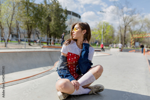 Outdoor portrait of charming stylish woman with long dark hair dressed in denim outfit and round yellow glasses sitting in the park in sunlight © PhotoBook