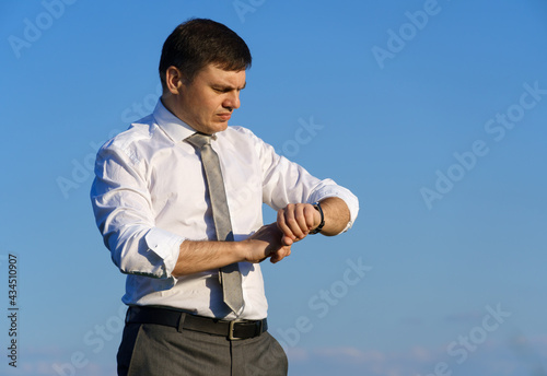 businessman posing in a field, he looks at his wristwatch, green grass and blue sky as background