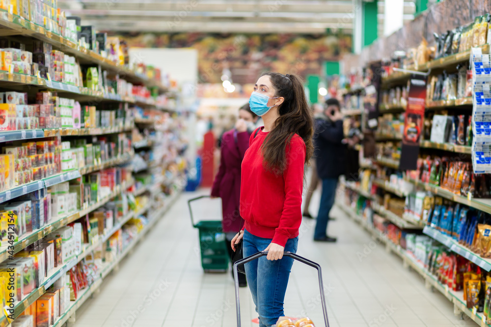 Shopping during the coronavirus pandemic. A young woman in a medical mask on her face looks at the windows with products in a supermarket. The concept of consumerism and the new reality