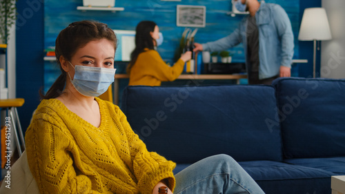 Lady looking at camera enoying spending time with friends in living room keeping social distancing wearing face mask to prevent coronavirus spreading. during global outbreak holding beer bottle. photo