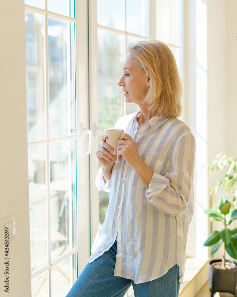 Enjoying new day. Side view of thoughtful beautiful mature woman holding cup of coffee or tea, looking in window and thinking or dreaming about something while spending free time in morning at home Enjoying new day. Side view of thoughtful beautiful mature woman holding cup of coffee or tea, looking in window and thinking or dreaming about something while spending free time in morning at home