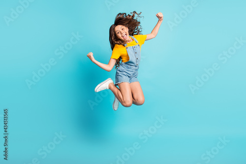 Full length photo of adorable strong small school girl wear denim jeans overall jumping showing muscles isolated blue color background