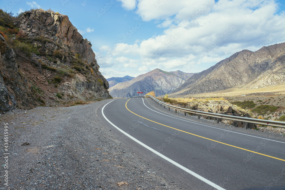 Colorful autumn landscape with mountain highway along rocks in sunshine. Bright alpine scenery with mountain road in autumn colors. Highway in mountains in fall time. Road along beautiful rocky wall. Colorful autumn landscape with mountain highway along rocks in sunshine. Bright alpine scenery with mountain road in autumn colors. Highway in mountains in fall time. Road along beautiful rocky wall.