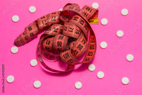 Measuring tape for measuring circumference of belly and hips lies on pink background surrounded by white pills in polka dot pattern. Concept photo of slimming for women with pills and pharmaceuticals