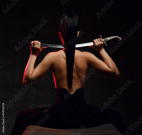Back view of young pretty brunette woman warrior in skirt and topless sitting back to camera holding sword on shoulders over dark background. Martial arts and beautiful women concept