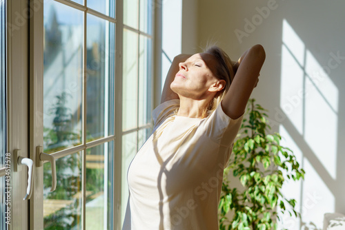 Enjoying new day. Beautiful happy mature woman stretching body and smiling while standing with closed eyes in sun rays shining through window on sunny morning at home. Optimism and happiness concept