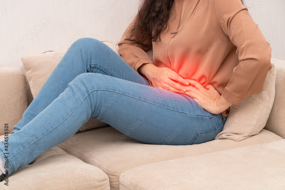 Unhappy Asian woman sitting on the sofa and holding on stomach suffering. Abdominal pain that comes from menstruation, diarrhea, or indigestion. Sickness and healthcare concept
