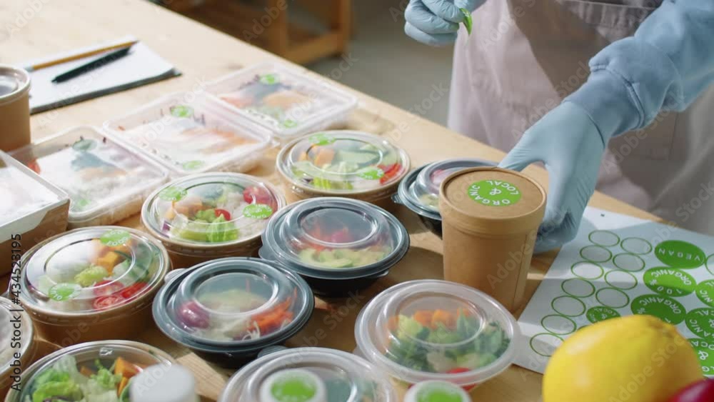 Zoom in shot if unrecognizable worker in apron and gloves putting stickers on ecofriendly containers with meals while working at healthy food delivery service Zoom in shot if unrecognizable worker in apron and gloves putting stickers on ecofriendly containers with meals while working at healthy food delivery service