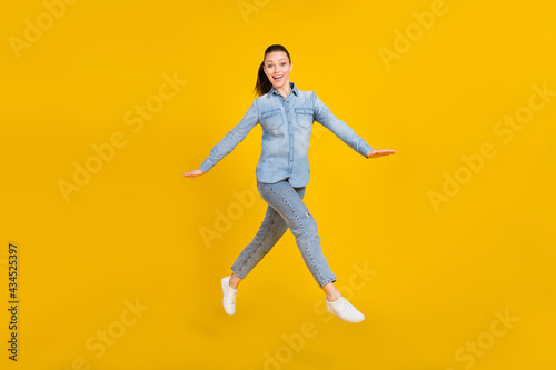 Full length photo of cute excited young woman wear denim shirt smiling jumping high isolated yellow color background