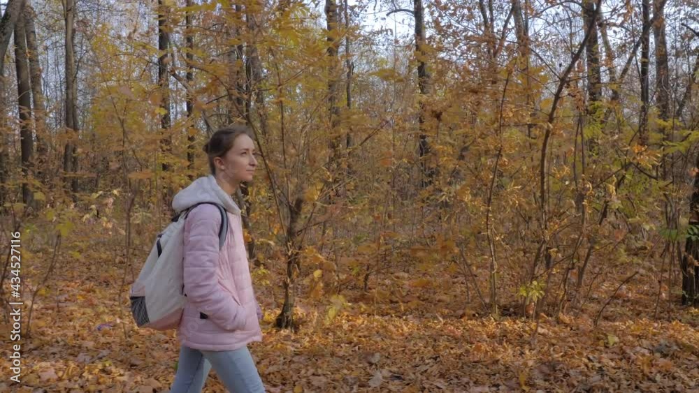 Side view of young woman in pink quilted jacket with backpack walking in autumn park, forest - wide angle steadicam shot. Active outdoor lifestyle, leisure time, freedom and adventure concept Side view of young woman in pink quilted jacket with backpack walking in autumn park, forest - wide angle steadicam shot. Active outdoor lifestyle, leisure time, freedom and adventure concept