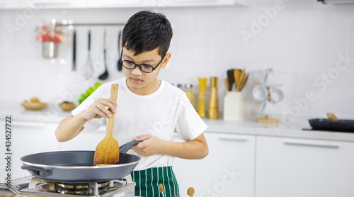 Little Asian boy in glasses frying patties on hot pan while preparing burgers for lunch in contemporary light kitchen at home. Concept for education and learning by doing.