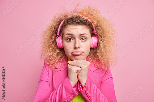 Frustrated European woman keeps hands under chin purses lips looks with sorrowful expression listens music via headphones dressed in bright clothes has bad mood isolated over pink background