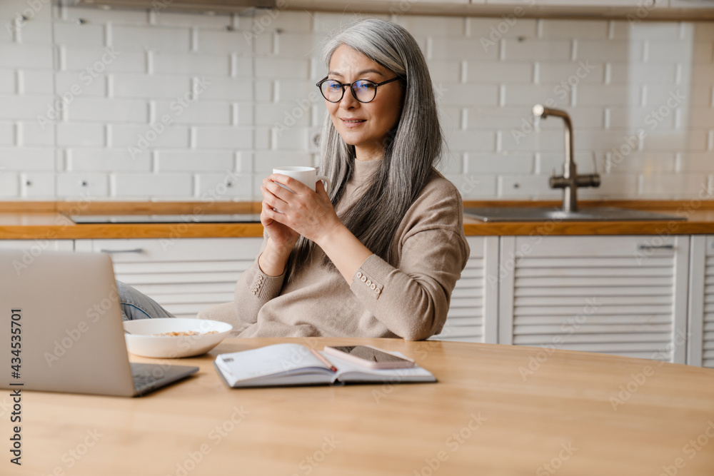 Mature smiling woman drinking coffee while working with laptop Mature smiling woman drinking coffee while working with laptop