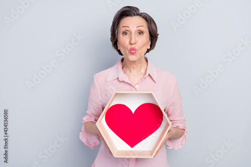 Portrait of attractive elderly middle-aged woman holding heart shape box sending air kiss isolated over gray pastel color background