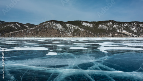 Deep intersecting cracks are visible on the frozen lake. Plots of snow on the surface of blue ice. On the shore there is a snow-covered wooded mountain range against the background of the sky. Baikal