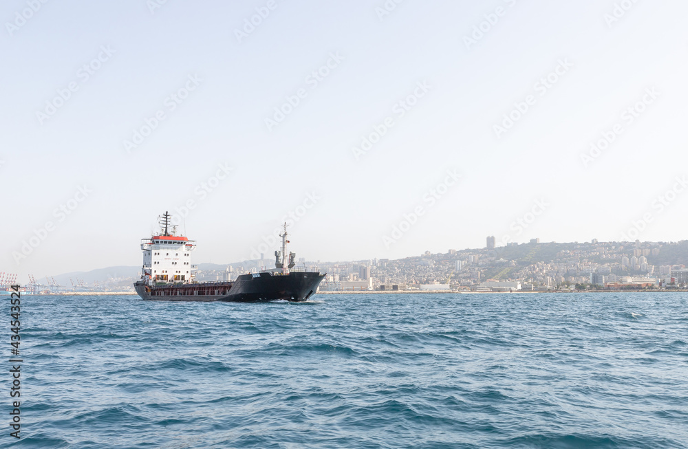 A large cargo ship is in the roadstead in the water area of the Haifa Bay, against the background of Haifa city, in the Mediterranean Sea, near the port of Haifa in Israel