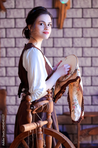 Portrait of Young Lovely Caucasian Brunette Woman Posing With Fancywork Hoop in Front of Spinning Wheel in Retro Dress In Rural Environment.