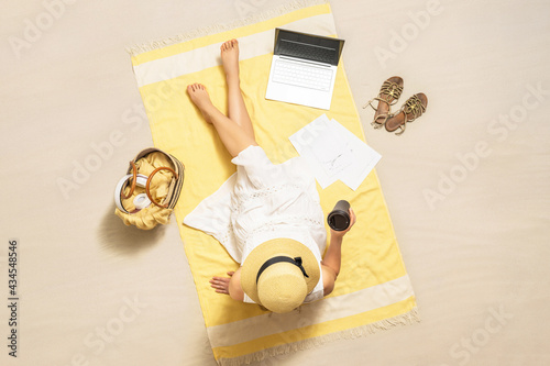Businesswoman freelancer drinking coffee sitting  on beach towel of sand beach and working on laptop with graphics and charts. Summer vacation. Aerial view