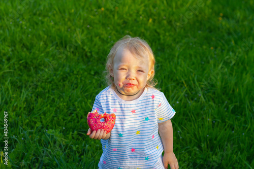 Picnic in the summer for children. Cute baby girl 2 years old eats a sweet pink donut in the summer in a green meadow