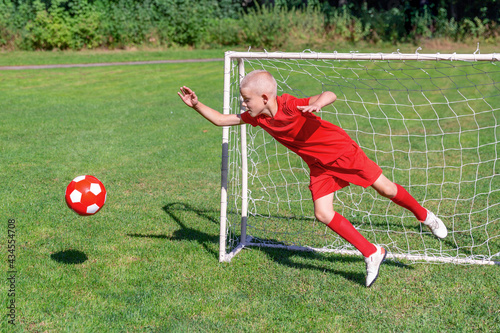 A young soccer player-goalkeeper in a red uniform catches the ball, defending the goal. Children's sports, physical activity concept