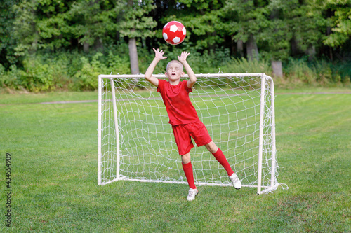 A young soccer player-goalkeeper in a red uniform catches the ball, defending the goal. Children's sports, physical activity concept