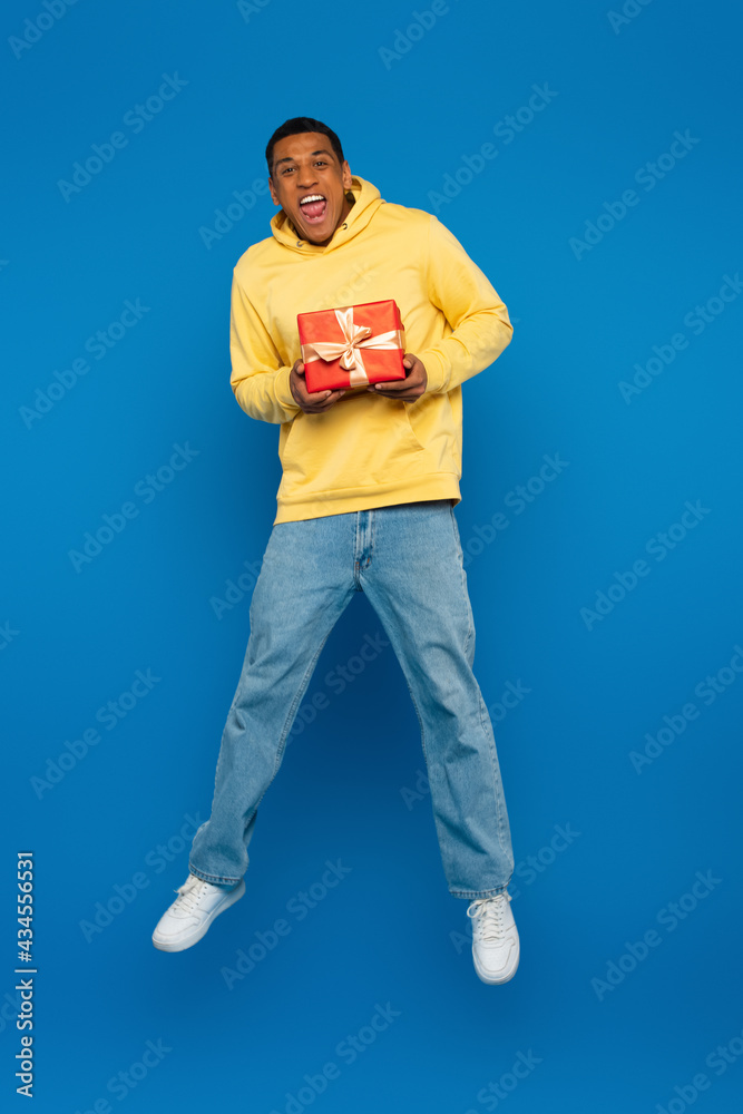 excited african american man levitating with red gift box on blue background excited african american man levitating with red gift box on blue background