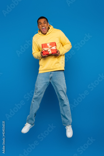 excited african american man levitating with red gift box on blue background