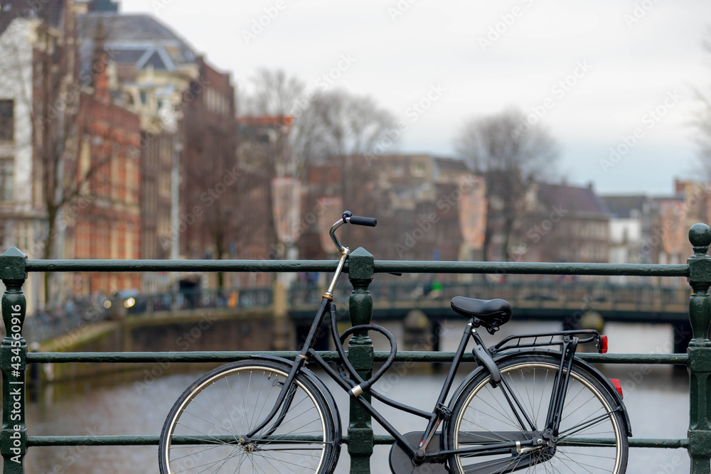 Selective focus of bicycle parked on canal bridge with blurred architecture features traditional houses as background, Amsterdam, Netherlands, Cycling is a common mode of transport in Holland.