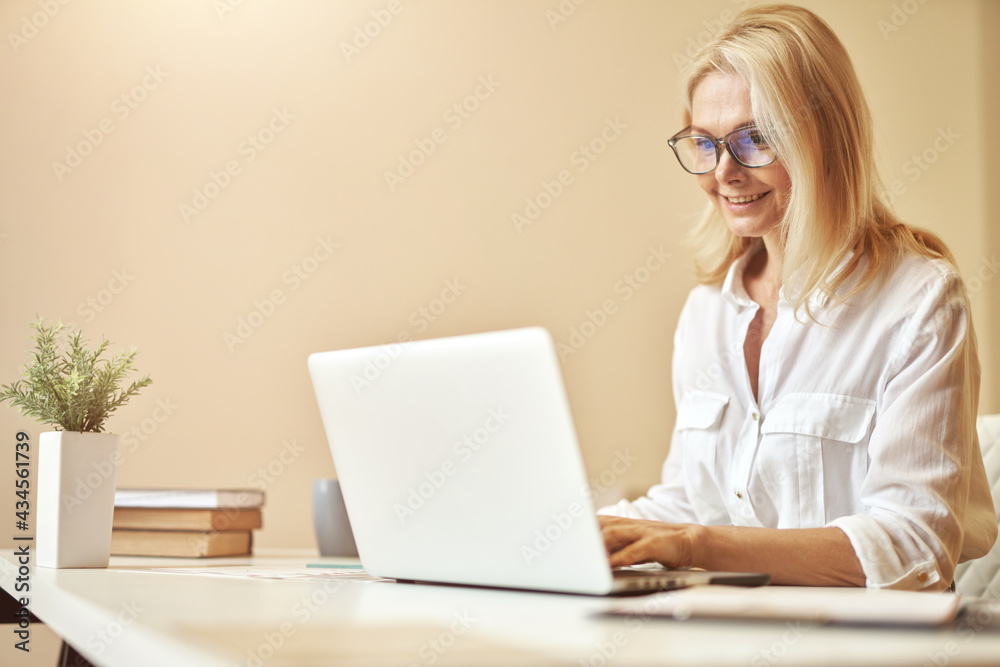 Happy mature businesswoman in glasses smiling while using laptop, working in the office Happy mature businesswoman in glasses smiling while using laptop, working in the office