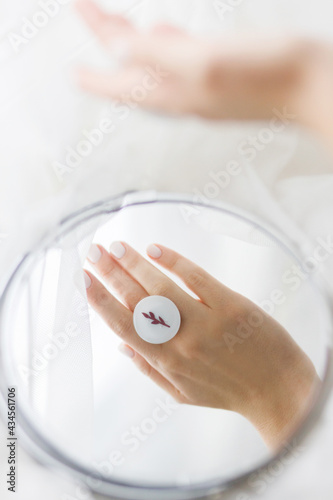 Stylish modern white round ring on beautiful hand reflected in mirror on background of soft white tulle. Unusual fashionable fused glass ring on female hand with white manicure.