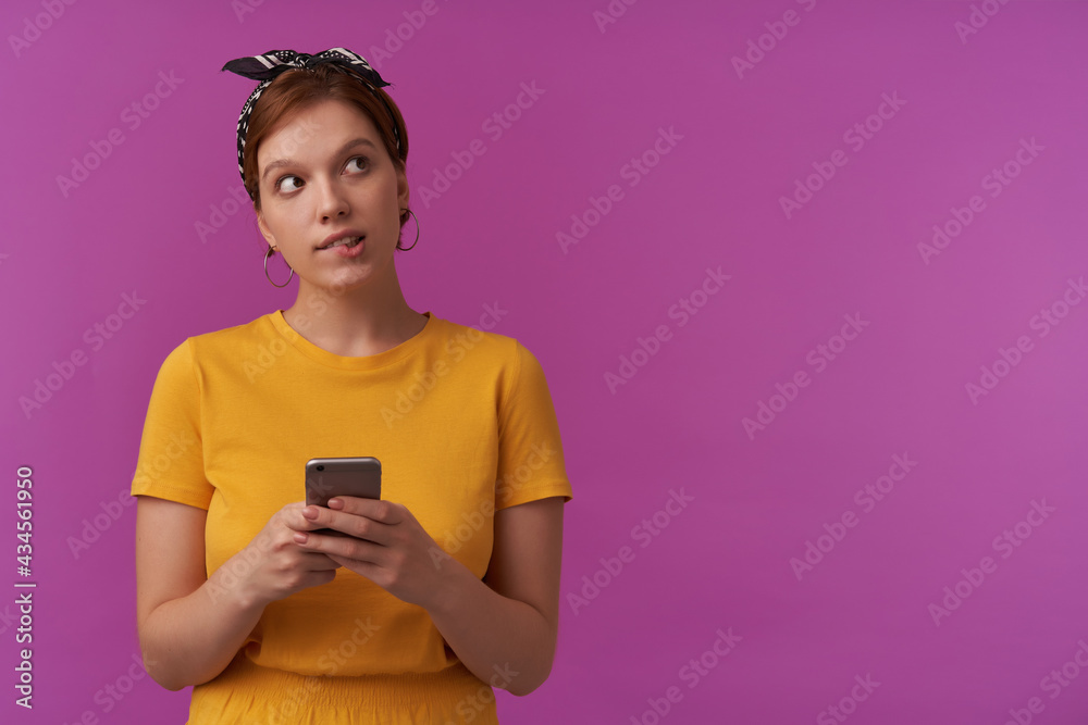 European charming woman 20s with natural makeup wearing yellow t-shirt and black bandana with arms hold phone emotion face looks aside dreaming counting calculating imagine against purple wall European charming woman 20s with natural makeup wearing yellow t-shirt and black bandana with arms hold phone emotion face looks aside dreaming counting calculating imagine against purple wall
