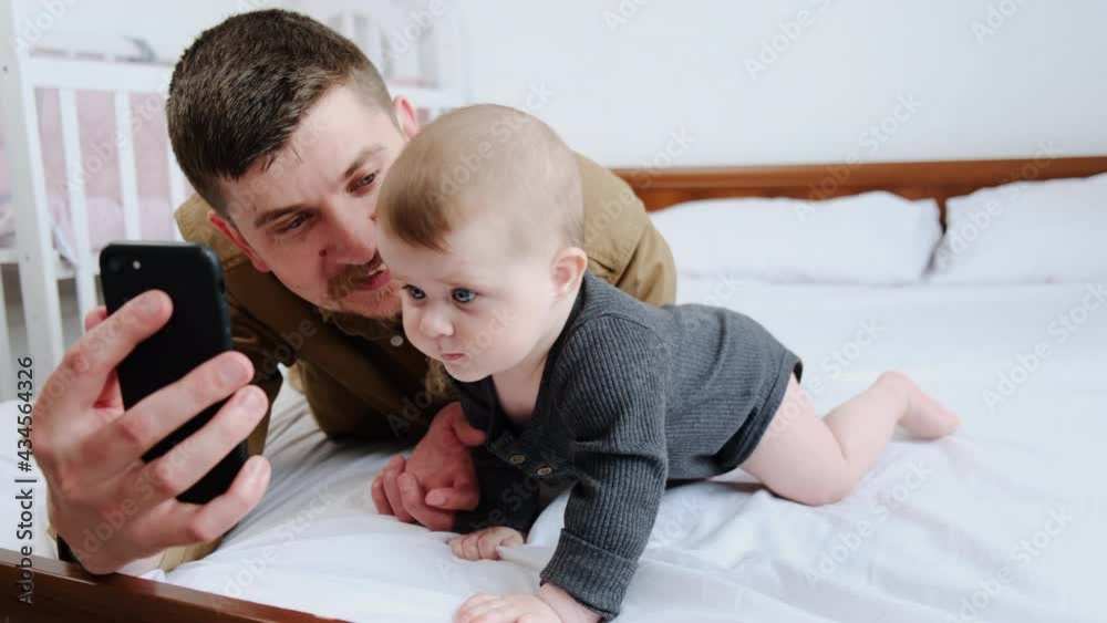 Close up of young father and cute beautiful newborn baby making selfie or video call to relatives lying on comfortable white bed at home. Technology, new generation, connection, parenthood concept Close up of young father and cute beautiful newborn baby making selfie or video call to relatives lying on comfortable white bed at home. Technology, new generation, connection, parenthood concept