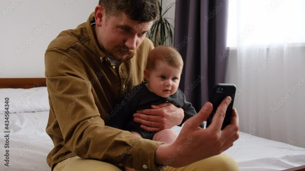 Happy young father and cute baby girl having video chat using smartphone, bearded dad sit on comfy bed with toddler enjoying mobile technology sharing fatherhood lifestyle with friend on social media Happy young father and cute baby girl having video chat using smartphone, bearded dad sit on comfy bed with toddler enjoying mobile technology sharing fatherhood lifestyle with friend on social media