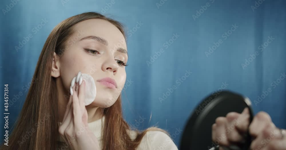Beautiful teenager washing off her makeup, wiping her face from cosmetic, using cotton pad, looking in mirror. Young woman posing in fashion studio on blue background. 
