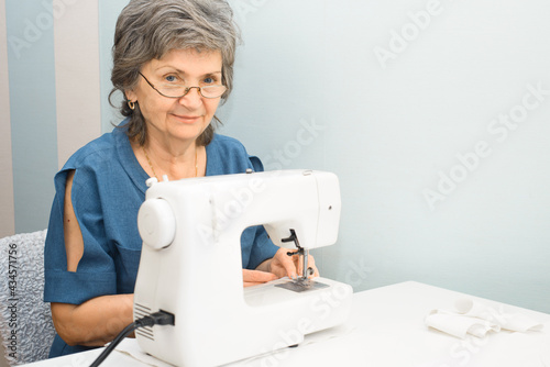 Portrait of smiling elderly woman with glasses using sewing machine at home. Senior woman sewing, indoors. Hobby or work at home for retired woman.