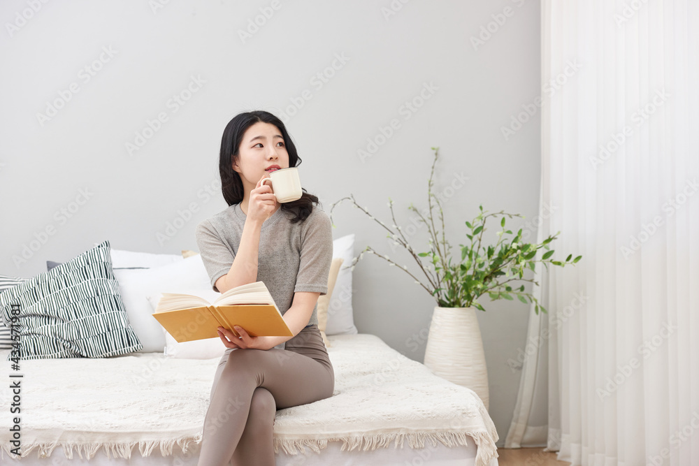 침실을 배경으로 침대에 앉아서 커피 또는 차를 마시며 책을 보고있는 젊은 아시아 한국 여자 모델
