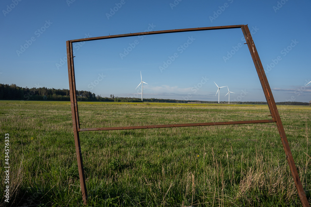 In the spring, a large rape field with a forest and blue skies behind it, there is a wind generator with a large propeller in the middle
