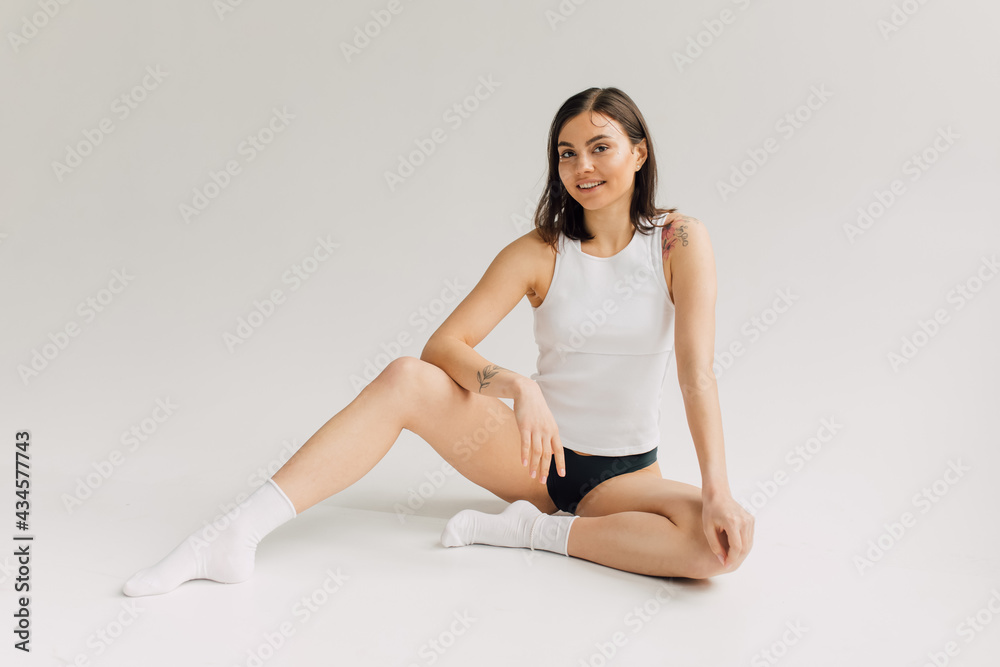 smiling young woman in white top, socks and black panties sitting on grey background