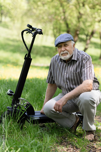 A positive, elderly man went for a walk to the park, stopped and sat down to rest next to an electric scooter. Summer, sunny day. Healthy lifestyle of the elderly.