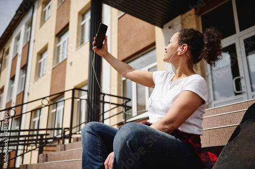 Young woman with ponytail and headphones in casual clothing making selfie while sitting on steps