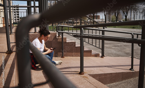 Young woman in headphones using mobile phone while sitting on skateboard on background of steps