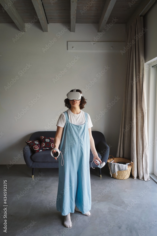 Female in modern VR goggles and with controllers standing in room at home and playing videogame in cyberspace