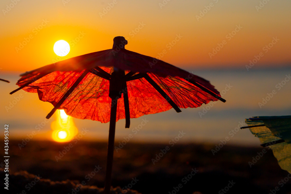 Paper cocktail umbrellas in sand on seashore at sunset dawn close-up.