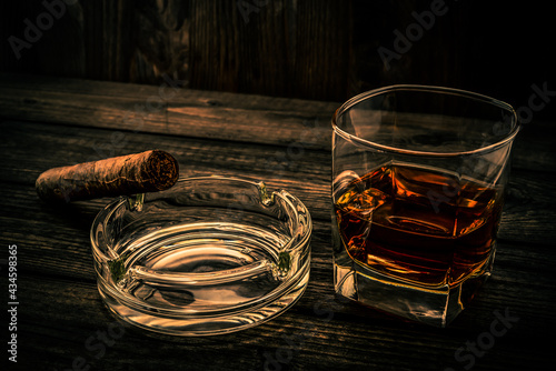Glass of brandy and cuban cigar with glass ashtray on an old wooden table. Angle view