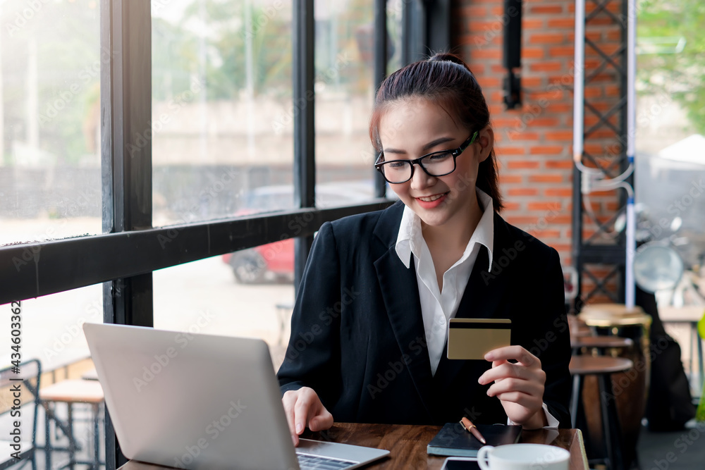 Young Asian businesswoman wearing glasses holding a credit card is shopping online using a laptop at the office.
