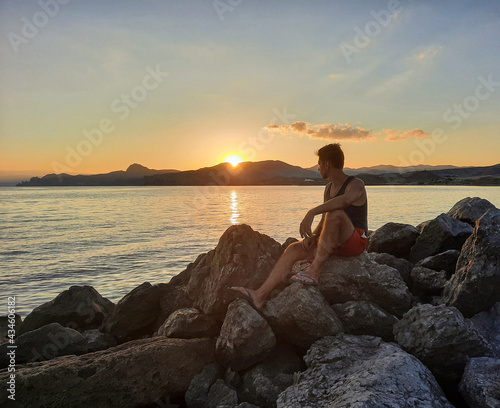 A man watches the sunset sitting on large rocks by the sea. A man enjoys the sunrise from behind the beautiful mountains. © OleJohny