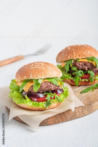 Принти на полотні Two juicy burgers made of grilled ground beef meat, cheese, lettuce, onion rings and tomatoes inside sliced bun with sesame served on cutting board on white wooden table with fork