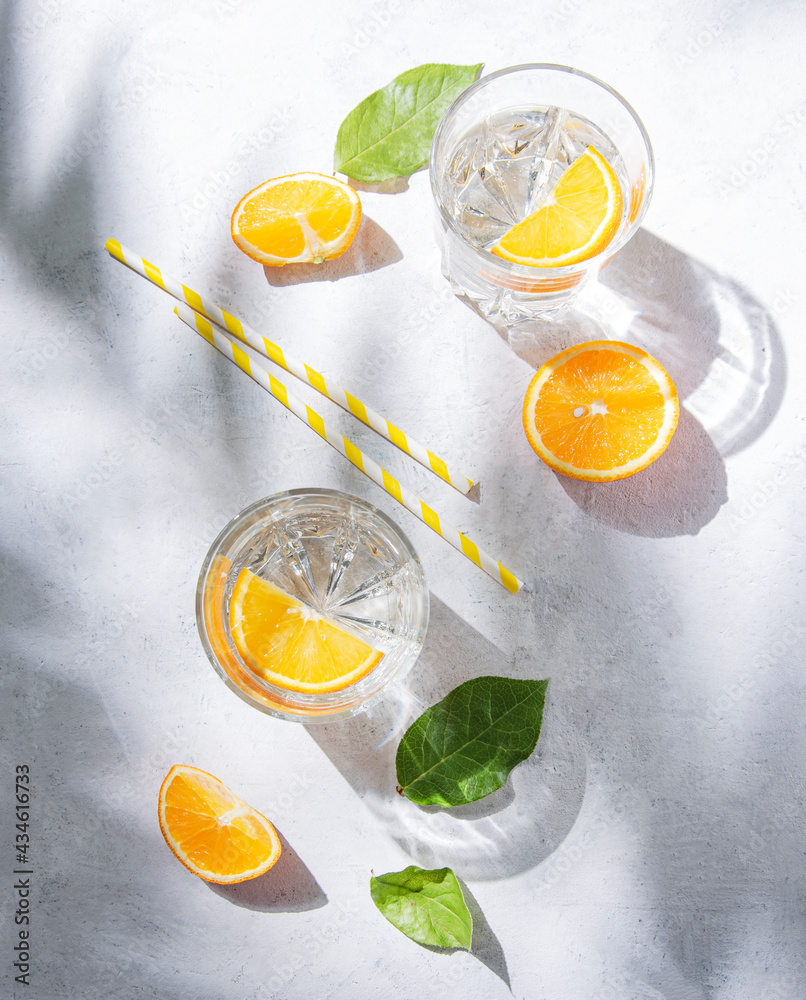 A glass of mineral water and a slice of orange on a gray table with fresh juicy fruit oranges with hard shadows. Summer time. Top view A glass of mineral water and a slice of orange on a gray table with fresh juicy fruit oranges with hard shadows. Summer time. Top view
