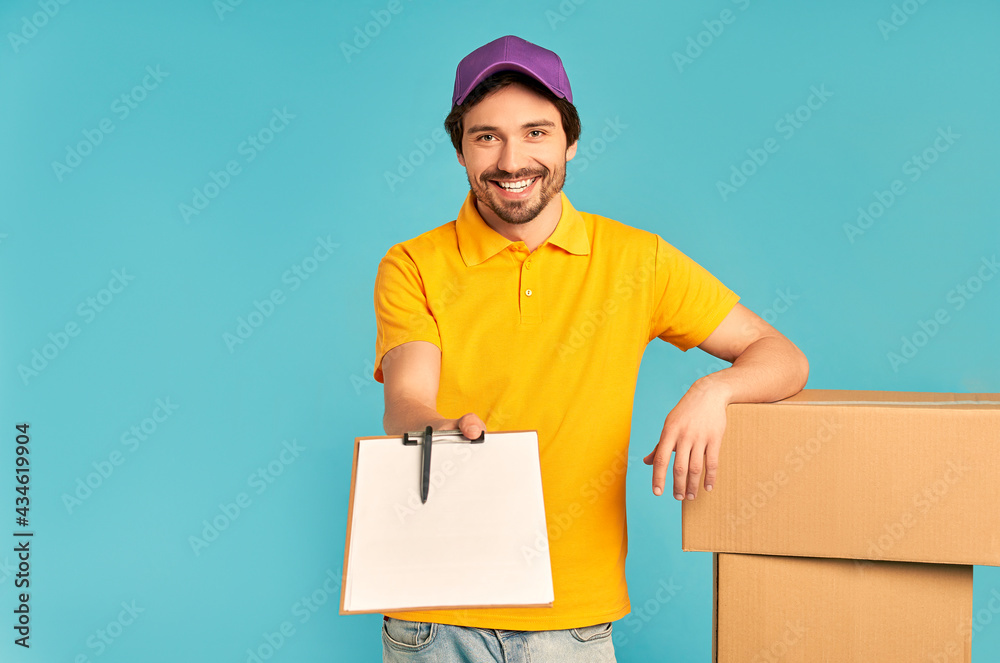 Young bearded courier delivery man in uniform with boxes holds out a document for signature isolated on blue background. Home delivery. Young bearded courier delivery man in uniform with boxes holds out a document for signature isolated on blue background. Home delivery.